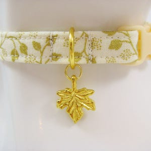 May include: A cream-colored pet collar with a gold floral pattern and a gold maple leaf charm. The collar has a cream-colored plastic buckle and a gold metal ring for the charm.