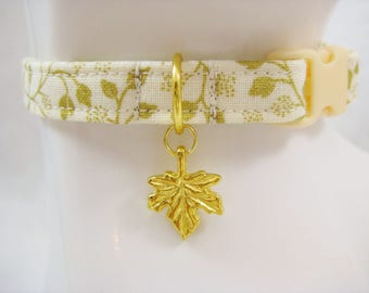 May include: A cream-colored pet collar with a gold floral pattern and a gold maple leaf charm. The collar has a cream-colored plastic buckle and a gold metal ring for the charm.