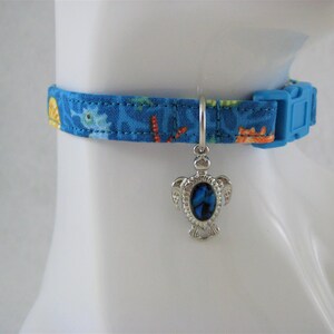 May include: Blue fabric pet collar with a colorful sea life pattern, featuring a blue plastic buckle. A silver-toned charm with a blue center hangs from the collar. The collar is designed to fit a small pet.