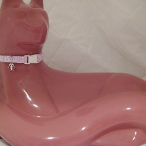 May include: A pink cat-shaped figurine wearing a light pink and white floral patterned cat collar with a white plastic buckle. The collar has a small charm with pink accents.