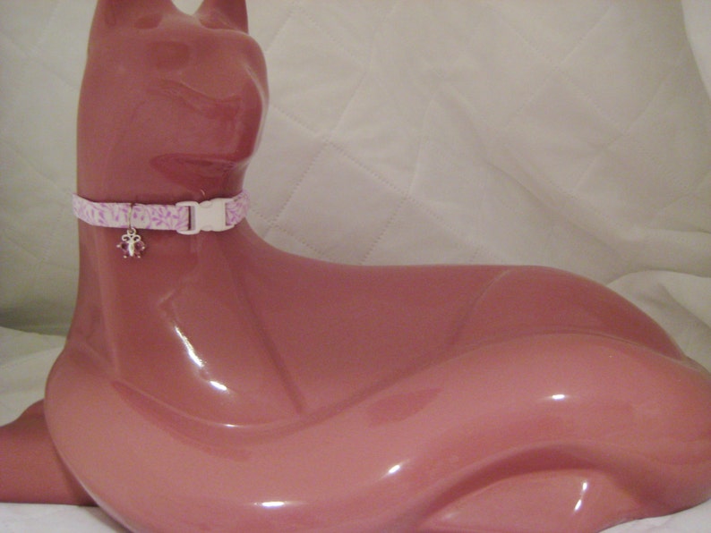 May include: A pink cat-shaped figurine wearing a light pink and white floral patterned cat collar with a white plastic buckle. The collar has a small charm with pink accents.