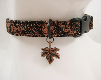 May include: A black and copper-colored dog collar with a leaf charm. The collar has a black plastic buckle and a copper-colored leaf-shaped charm. The collar is made of a patterned fabric with copper and black speckles.