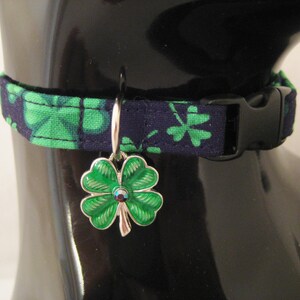 May include: A navy blue and green patterned pet collar with a black buckle. The collar features a silver charm with a green four-leaf clover design and a small jewel in the center. The collar is displayed on a black mannequin.
