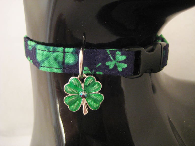 May include: A navy blue and green patterned pet collar with a black buckle. The collar features a silver charm with a green four-leaf clover design and a small jewel in the center. The collar is displayed on a black mannequin.