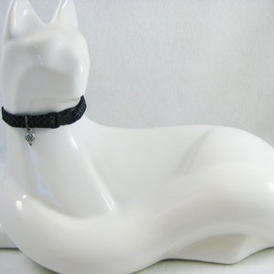 May include: A black cat collar with a diamond pattern and a silver charm. The collar is secured with a black plastic buckle. The collar is displayed on a white cat-shaped figurine.