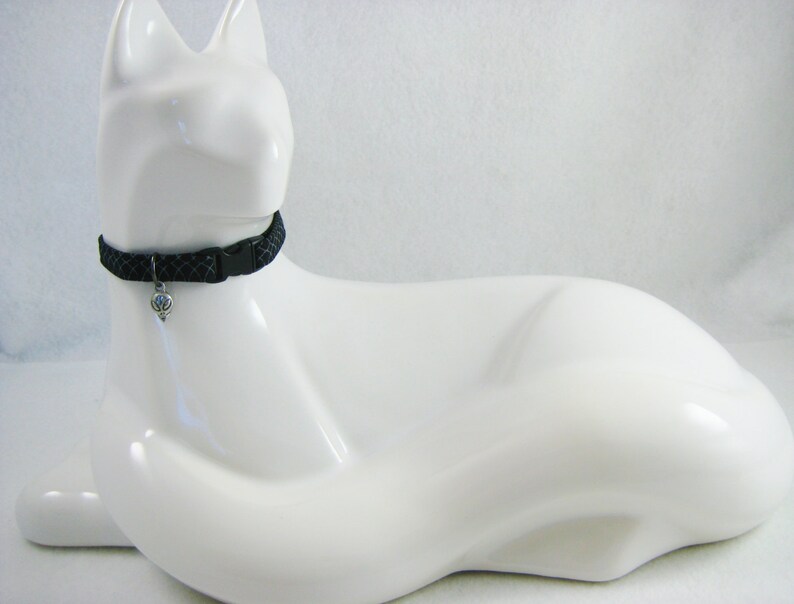 May include: A black cat collar with a diamond pattern and a silver charm. The collar is secured with a black plastic buckle. The collar is displayed on a white cat-shaped figurine.