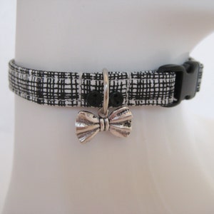May include: A black and white plaid cat collar with a silver bow charm. The collar has a black plastic buckle and a silver metal loop for attaching tags. The collar is displayed on a white mannequin head.
