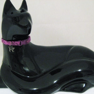 May include: A glossy black cat figurine with a purple collar featuring a floral pattern and a silver cat charm. The cat is in a reclining pose, with its ears perked up.