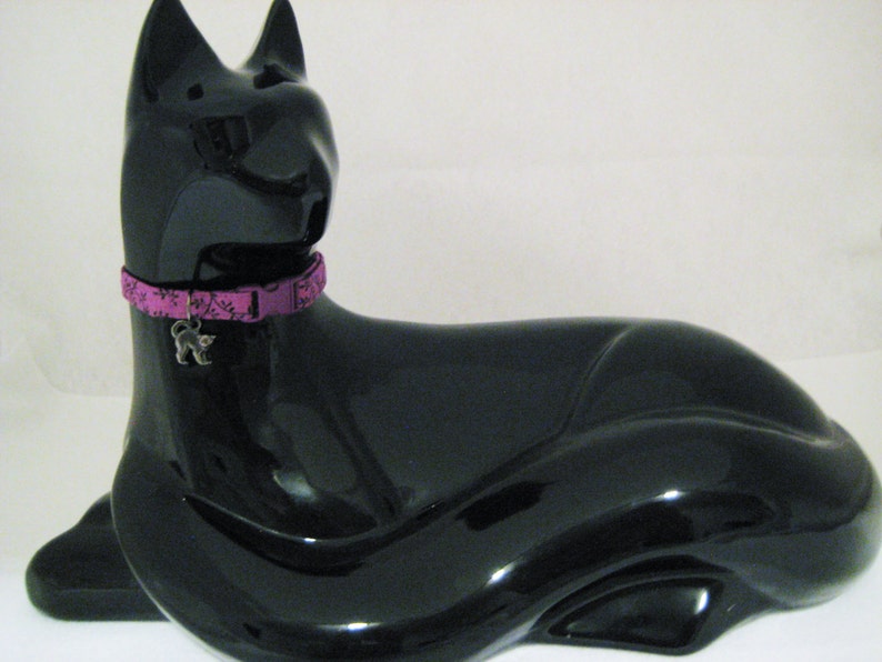 May include: A glossy black cat figurine with a purple collar featuring a floral pattern and a silver cat charm. The cat is in a reclining pose, with its ears perked up.