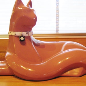 K&ouml;nnte beinhalten: Gl&auml;nzende, terrakottafarbene Katzenfigur in entspannter Pose. Die Katze tr&auml;gt ein wei&szlig;es Halsband mit schwarzen Pfotenabdr&uuml;cken und einem kleinen Anh&auml;nger. Die Figur steht auf einer Holzoberfl&auml;che, mit einem Fenster im Hintergrund.