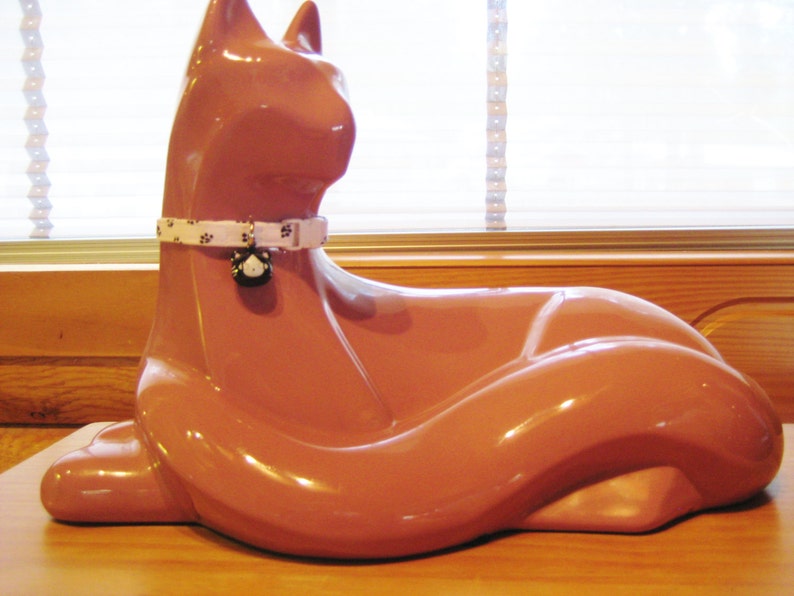 K&ouml;nnte beinhalten: Gl&auml;nzende, terrakottafarbene Katzenfigur in entspannter Pose. Die Katze tr&auml;gt ein wei&szlig;es Halsband mit schwarzen Pfotenabdr&uuml;cken und einem kleinen Anh&auml;nger. Die Figur steht auf einer Holzoberfl&auml;che, mit einem Fenster im Hintergrund.