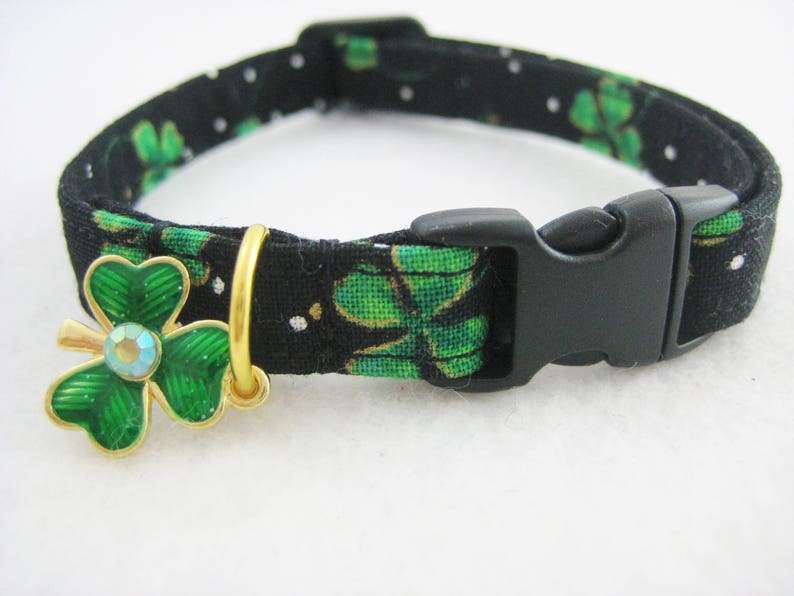May include: Black cat collar with a green shamrock pattern and a gold-tone shamrock charm with a blue crystal center. The collar has a black plastic buckle.