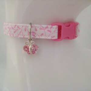 May include: A pink and white patterned cat collar with a pink plastic buckle. The collar features a silver charm in the shape of a butterfly with pink gemstones. The collar is designed to fit a cat's neck.