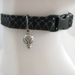 May include: Black fabric collar with a geometric pattern and a silver alien head charm. The collar has a black plastic buckle. The charm is suspended from a silver ring.