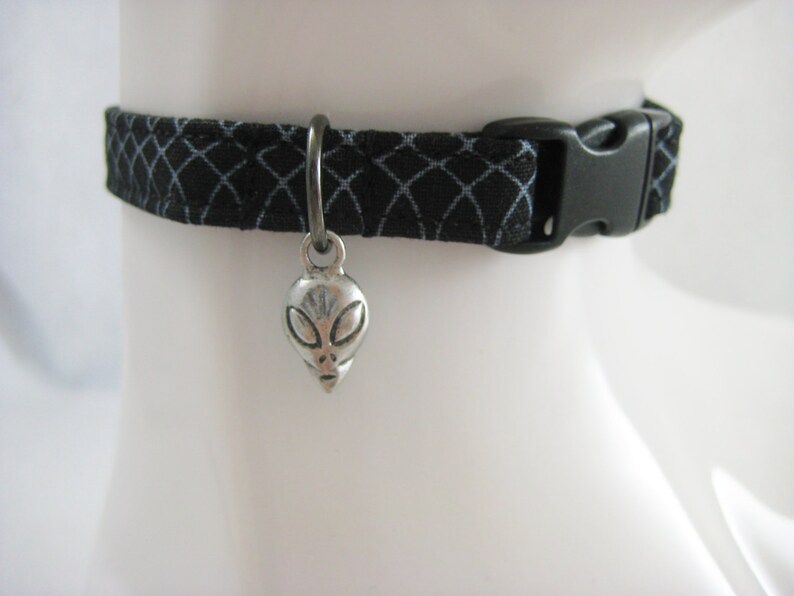 May include: Black fabric collar with a geometric pattern and a silver alien head charm. The collar has a black plastic buckle. The charm is suspended from a silver ring.
