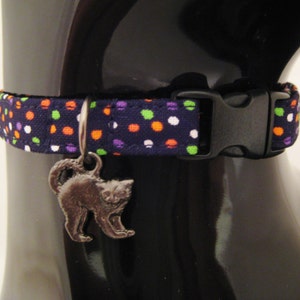 May include: A navy blue cat collar with a repeating pattern of orange, green, purple, and white polka dots. The collar has a black plastic buckle and a silver cat charm. This pet accessory is designed for cats.