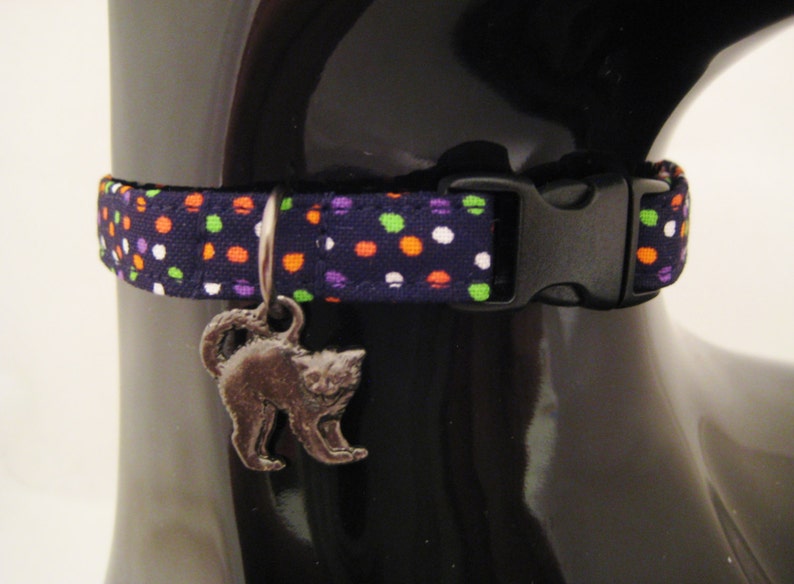 May include: A navy blue cat collar with a repeating pattern of orange, green, purple, and white polka dots. The collar has a black plastic buckle and a silver cat charm. This pet accessory is designed for cats.