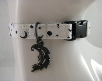 May include: White cat collar with a black crescent moon and dot pattern. The collar has a black plastic buckle and a dark metal charm of a crescent moon with a face. The collar is designed for a cat.