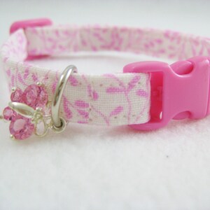 May include: A pink and white floral patterned pet collar with a pink plastic buckle. The collar features a silver charm with pink crystal accents. The collar is designed for small pets.