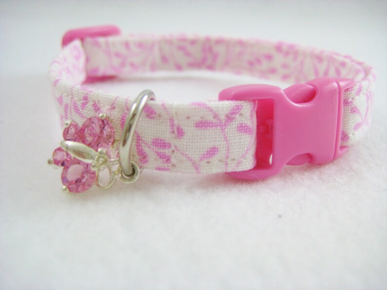May include: A pink and white floral patterned pet collar with a pink plastic buckle. The collar features a silver charm with pink crystal accents. The collar is designed for small pets.