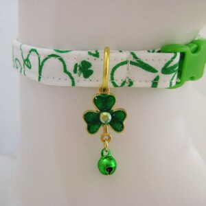 May include: A white cat collar with a green clover pattern and a green plastic buckle. The collar features a gold-colored charm with a green shamrock design and a small green bell.