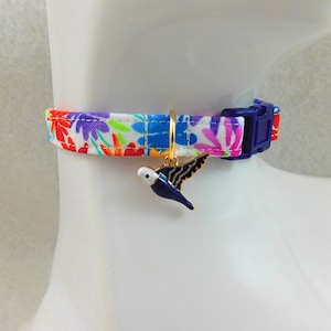 May include: A colorful cat collar with a floral pattern in red, purple, blue, and green. The collar has a blue plastic buckle and a gold-colored charm of a blue and white bird with black wings.