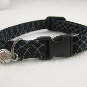May include: Black cat collar with a white geometric pattern. The collar features a black plastic buckle and a silver alien charm. Designed for a cat.