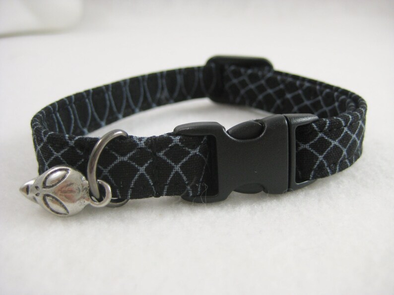 May include: Black cat collar with a white geometric pattern. The collar features a black plastic buckle and a silver alien charm. Designed for a cat.