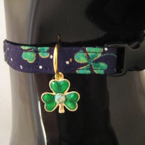May include: A navy blue pet collar with a green shamrock pattern and a black plastic buckle. A gold-colored charm with a green shamrock design and a central crystal hangs from the collar, adding a touch of elegance.