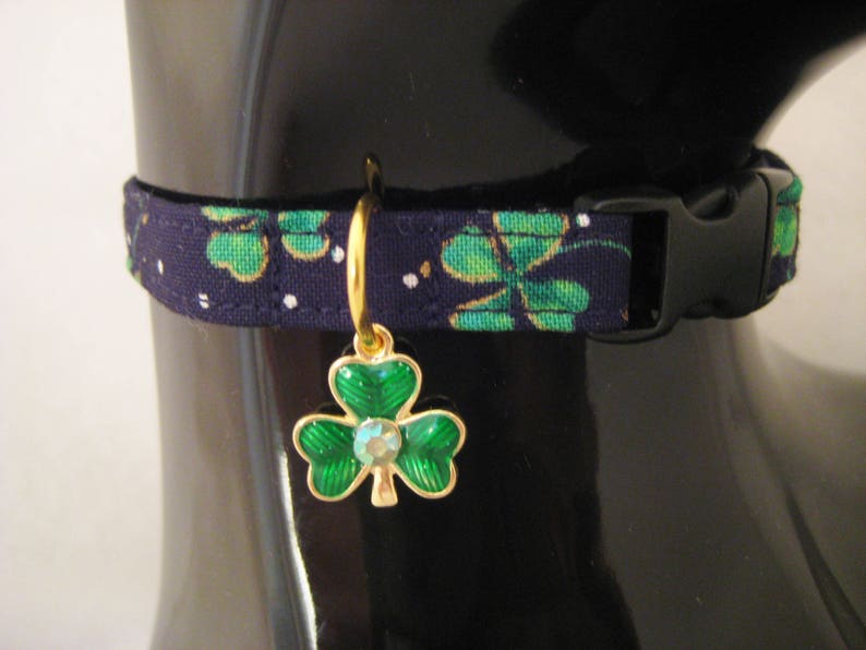 May include: A navy blue pet collar with a green shamrock pattern and a black plastic buckle. A gold-colored charm with a green shamrock design and a central crystal hangs from the collar, adding a touch of elegance.