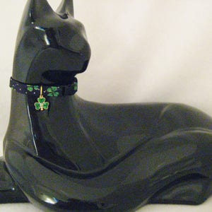 May include: A glossy black cat figurine wearing a patterned collar with green shamrocks and a gold charm. The charm features a green shamrock with a small clear gem in the center. The collar is dark blue with green shamrocks and small gold dots.