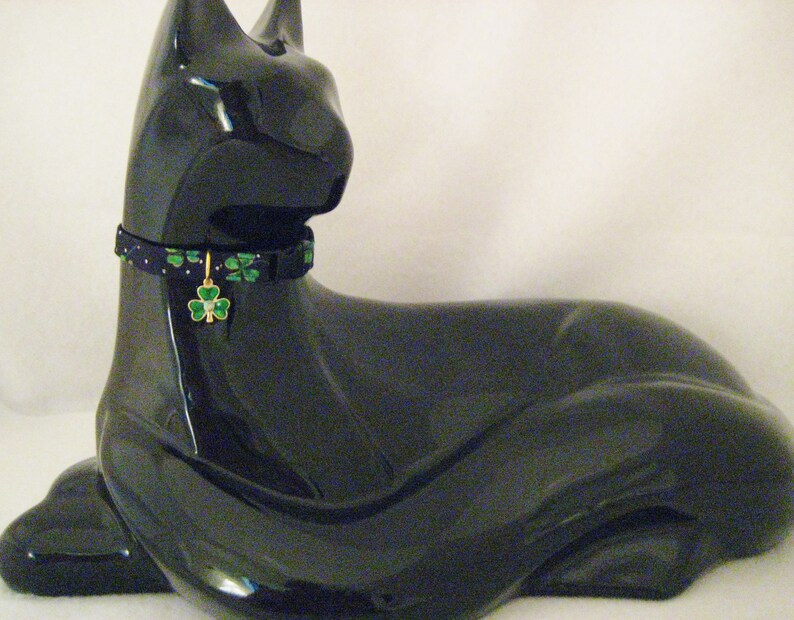 May include: A glossy black cat figurine wearing a patterned collar with green shamrocks and a gold charm. The charm features a green shamrock with a small clear gem in the center. The collar is dark blue with green shamrocks and small gold dots.