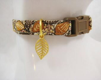 May include: A patterned cat collar with a gold leaf charm. The collar features a brown plastic buckle and a fabric design with orange and brown leaf patterns. The gold leaf charm hangs from a gold ring.