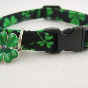 May include: A black and green cat collar with a clover pattern. The collar has a black plastic buckle and a silver charm with a green clover design and a small, iridescent gem in the center.