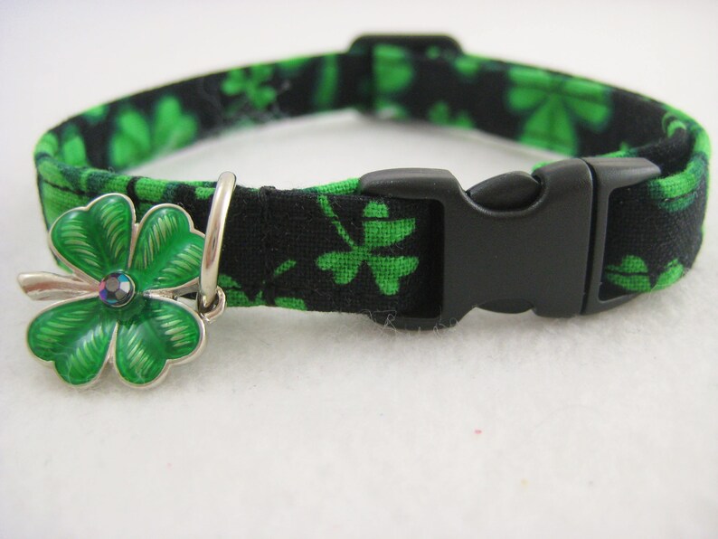 May include: A black and green cat collar with a clover pattern. The collar has a black plastic buckle and a silver charm with a green clover design and a small, iridescent gem in the center.