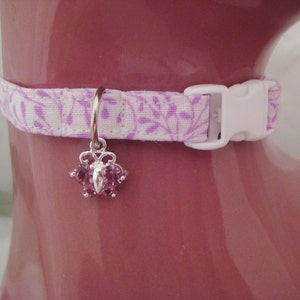 May include: A white and lavender floral patterned cat collar with a white plastic buckle. A silver charm with pink gemstones hangs from the collar. The collar is displayed on a pink surface.