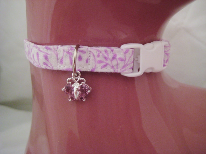 May include: A white and lavender floral patterned cat collar with a white plastic buckle. A silver charm with pink gemstones hangs from the collar. The collar is displayed on a pink surface.