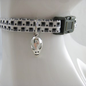 May include: A pet collar featuring a monochrome geometric design. It has a silver alien charm and a dark green buckle. The collar is designed to fit around a pet's neck.