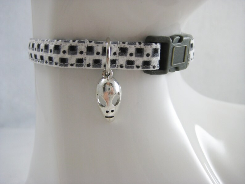 May include: A pet collar featuring a monochrome geometric design. It has a silver alien charm and a dark green buckle. The collar is designed to fit around a pet's neck.