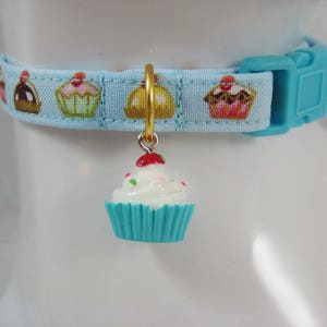 May include: A light blue pet collar featuring a cupcake design. The collar has a turquoise plastic buckle and a gold-coloured charm with a cupcake pendant. The cupcake charm has white frosting with sprinkles and a red cherry.