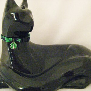 May include: A glossy black cat figurine with a green and black patterned collar and a shamrock charm. The cat is in a resting pose, with its paws extended. The figurine is a decorative item.