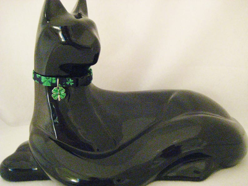 May include: A glossy black cat figurine with a green and black patterned collar and a shamrock charm. The cat is in a resting pose, with its paws extended. The figurine is a decorative item.