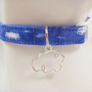 May include: Blue fabric pet collar with a white cloud pattern and a silver cloud charm. The collar has a white plastic buckle. The charm is a cloud shape. The collar is around a neck.