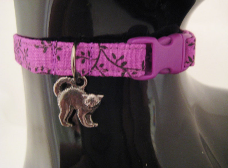 May include: A purple cat collar with a black floral pattern and a matching purple buckle. A silver cat charm hangs from the collar. The collar is designed for a cat.