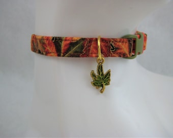 May include: A fall-themed cat collar with a leaf charm. The collar features a pattern of orange, brown, and green leaves. A gold-colored leaf charm hangs from the collar. The collar has a green plastic buckle.