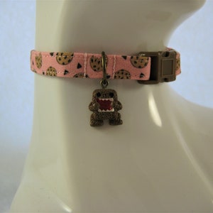 May include: A pink pet collar with a repeating pattern of chocolate chip cookies. The collar has a brown plastic buckle and a bronze-colored charm of a cartoon monster with a red mouth. The collar is displayed on a white mannequin.