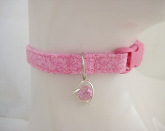 May include: Pink pet collar with a floral pattern and a pink plastic buckle. A silver charm with a pink heart-shaped gem hangs from the collar. The collar is designed to fit around a pet's neck.