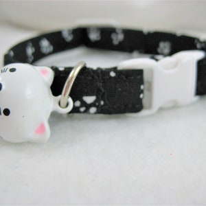 K&ouml;nnte beinhalten: Schwarzes Katzenhalsband mit wei&szlig;en Pfotenabdr&uuml;cken und wei&szlig;er Schnalle. Das Halsband hat eine wei&szlig;e, katzenf&ouml;rmige Glocke mit rosa Ohren und schwarzen Gesichtsdetails. F&uuml;r Haustiere.