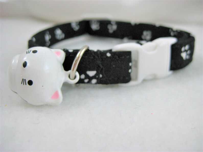 K&ouml;nnte beinhalten: Schwarzes Katzenhalsband mit wei&szlig;en Pfotenabdr&uuml;cken und wei&szlig;er Schnalle. Das Halsband hat eine wei&szlig;e, katzenf&ouml;rmige Glocke mit rosa Ohren und schwarzen Gesichtsdetails. F&uuml;r Haustiere.