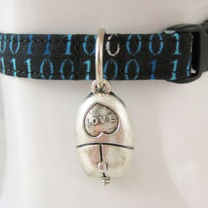May include: Black cat collar with a binary code pattern in blue and white. The collar has a black plastic buckle and a silver-colored charm with the word "LOVE" inside a heart.